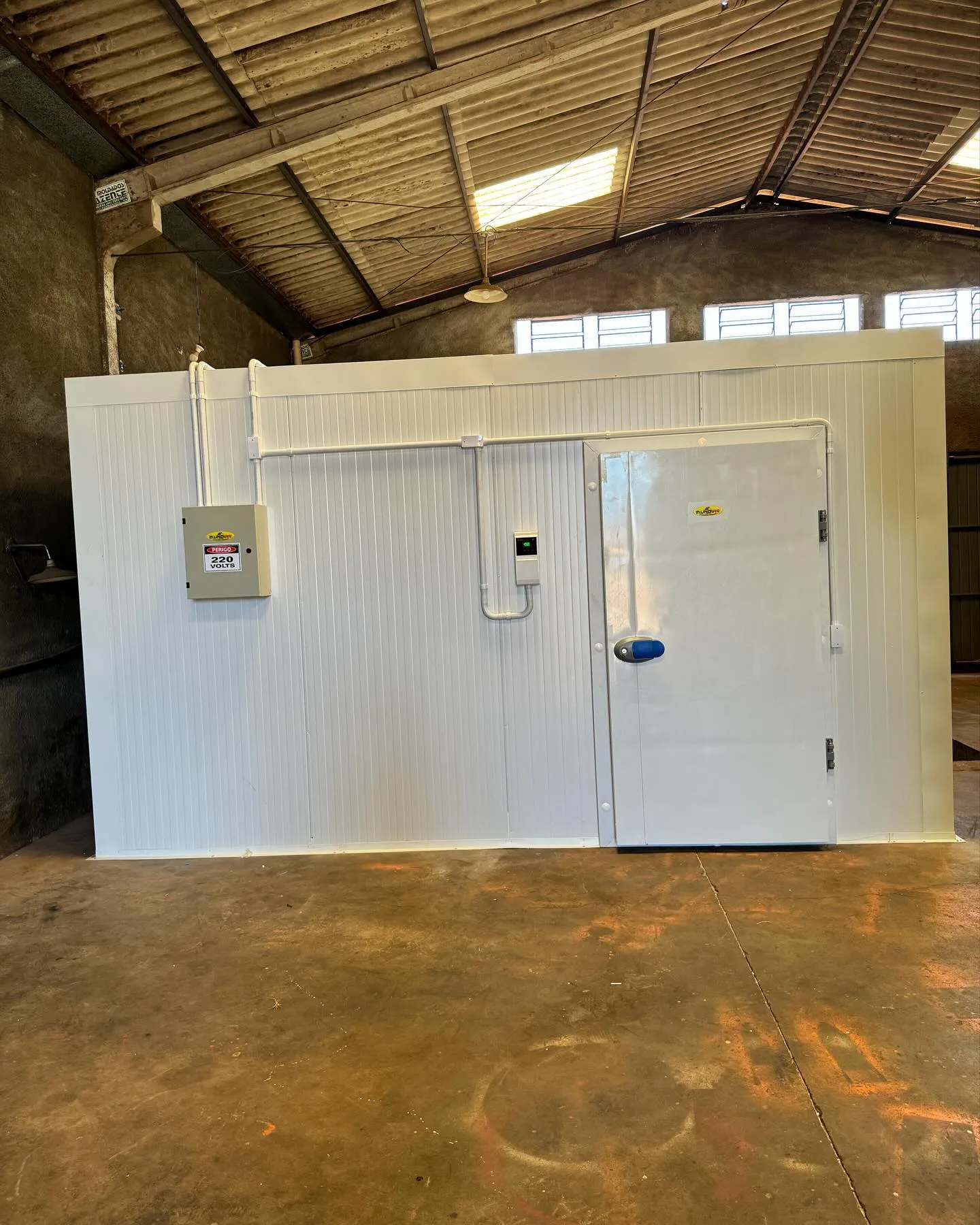Câmara fria modular branca instalada em galpão, com porta, quadro elétrico e controlador