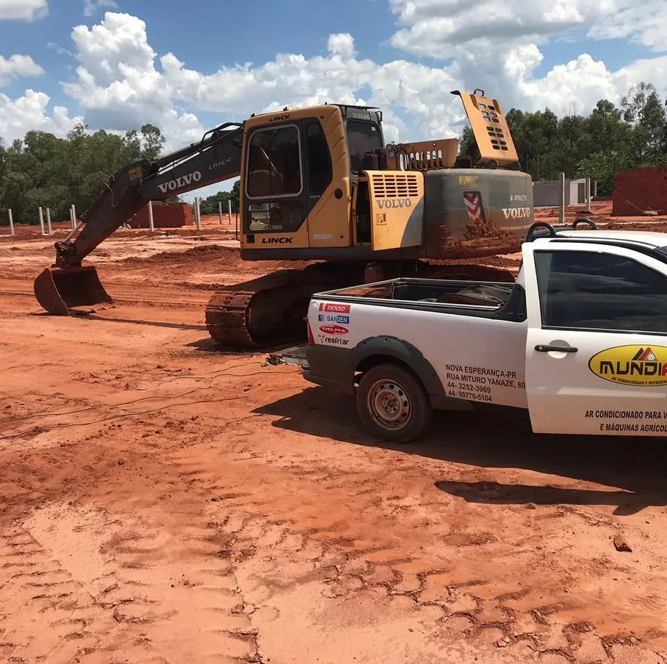 Escavadeira Volvo em canteiro de terra vermelha com picape de atendimento Mundiar